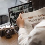 Jurnalistă care citește un comunicat de presă pe laptop, într-o redacție cu birouri și monitoare